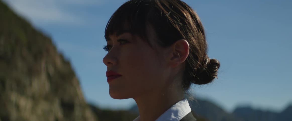 Woman with her dark hair in a bun stands near the mountains looking pensive
