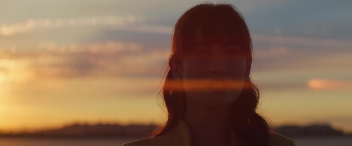 A young woman stands before a setting sun while looking off into the distance