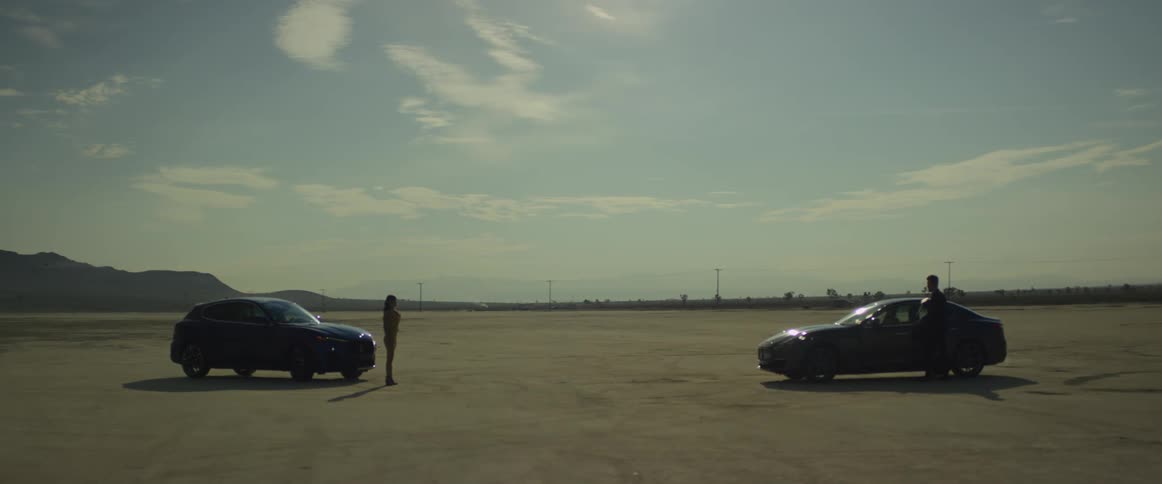 Two automobiles are parked facing each other and a woman is standing in front of one and a man gets out of the car and walks toward the woman