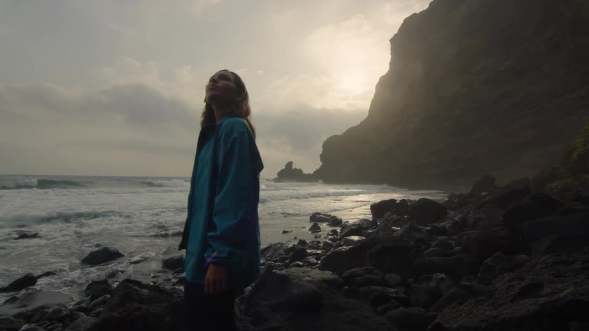 Woman with wavy hair and blue raincoat stands on the beach at sunset looking around
