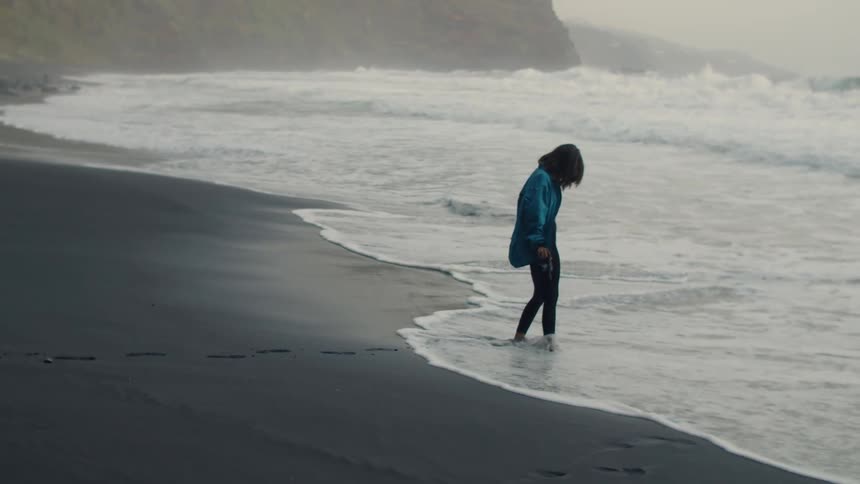 A woman walking barefoot on the beach towards the waves lifts her camera and takes a photo