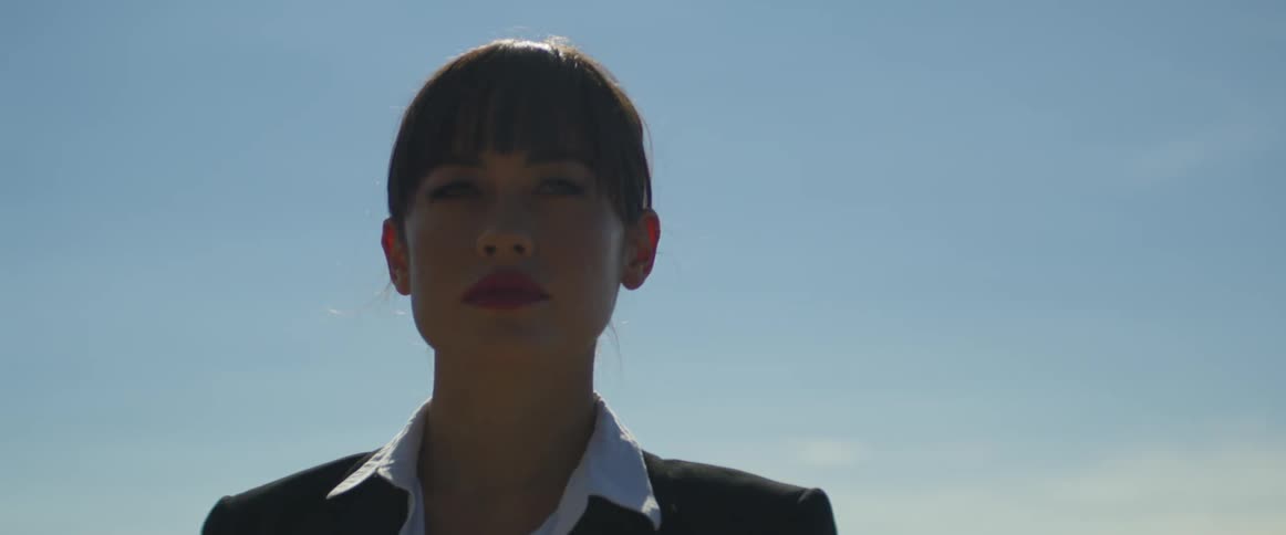 A woman with hair in a bun and wearing business attire stands outside in the sunlight