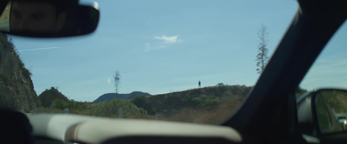 A man drives a car on a mountain road and the silhouette of a man standing on a ridge is visible