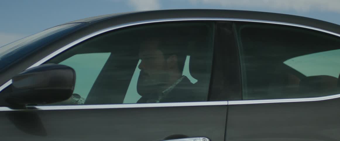 A man is seen through the drivers side window driving a grey automobile traveling on a road