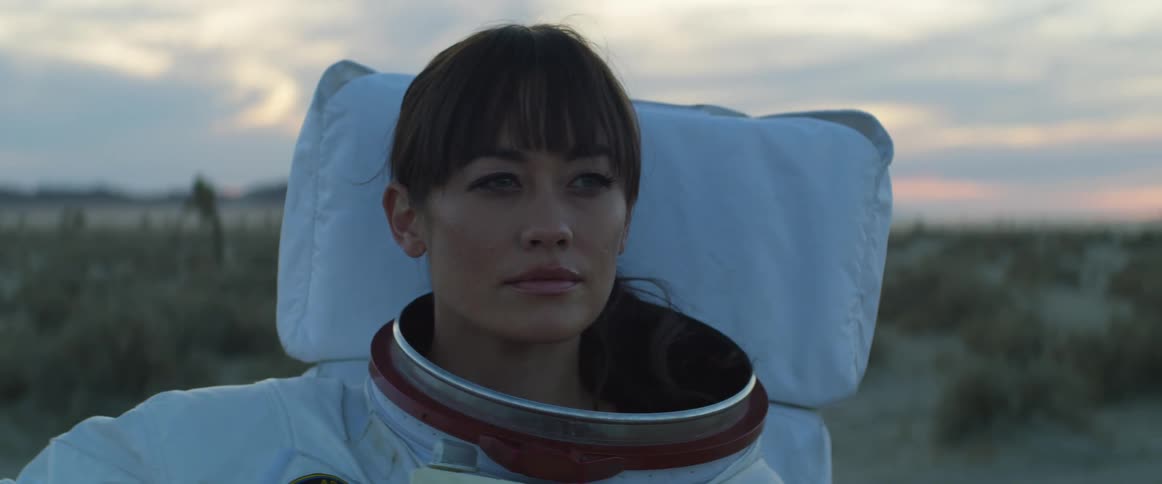 Young woman wearing an astronaut suit stands in the desert at sunset looking at the horizon