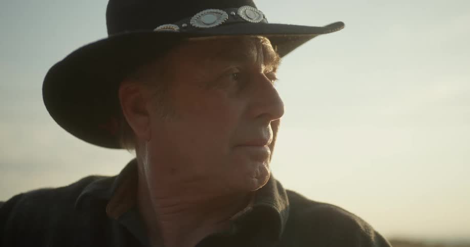 An older white man wearing a cowboy hat is sitting outside
