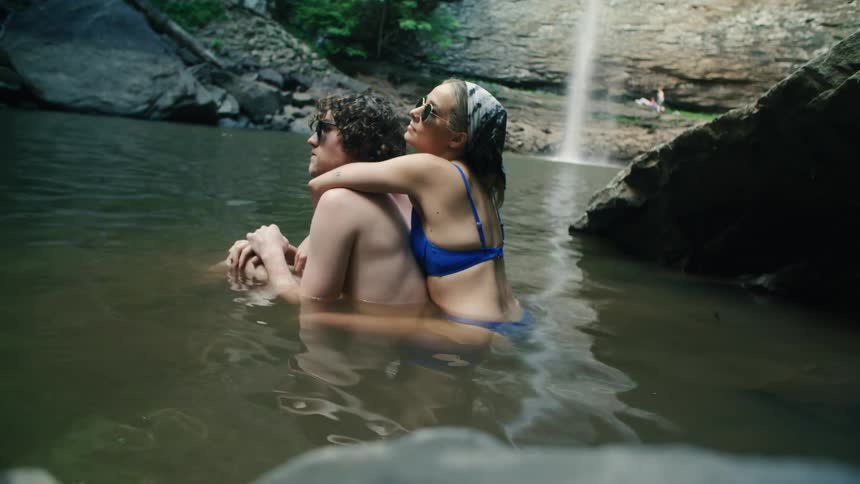 A man and woman are in the water near a waterfall and she is riding on him piggy back style with her arms around his neck