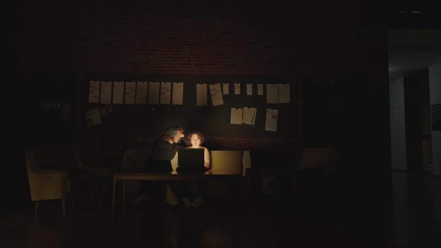 Two people are sitting on a couch in a dark room with a laptop computer on the table in front of them