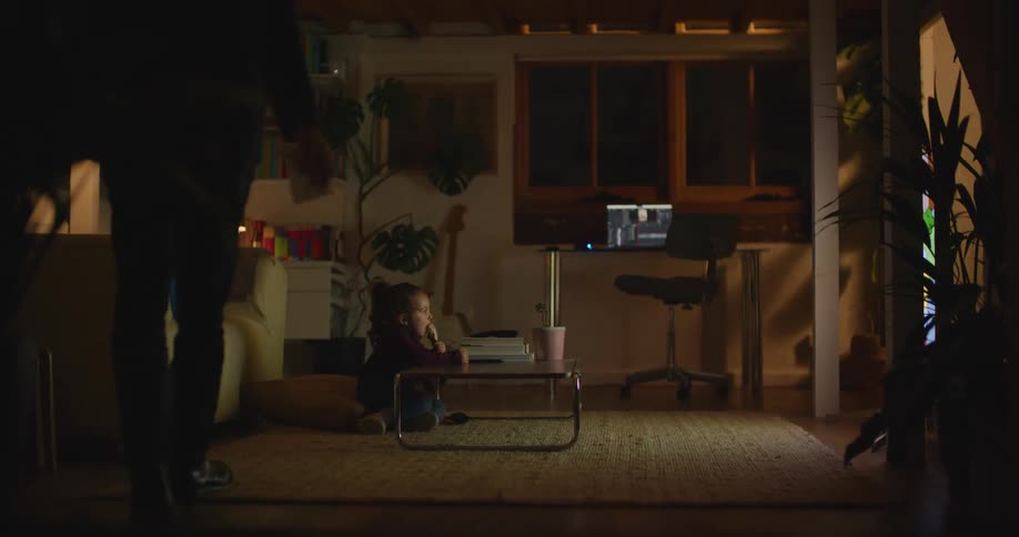 Man walks into the living room and sits on the floor next to his daughter and they watch television and eat ice cream