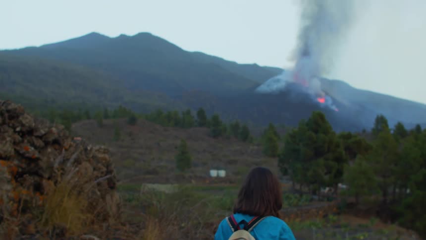 A woman wearing a backpack is walking on a mountain road and there is smoke and fire on a mountain in the distance
