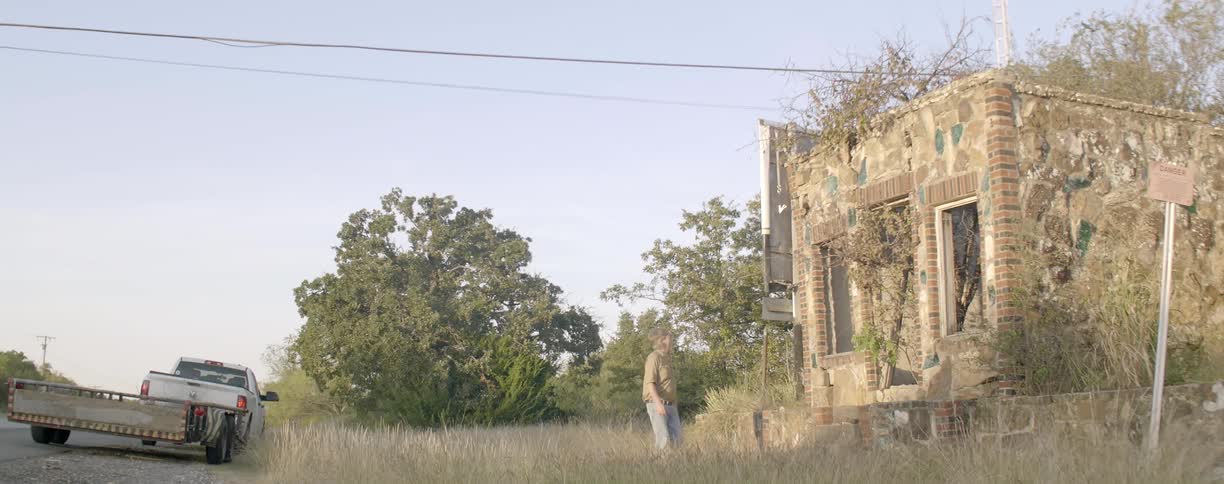 An older man approaches a run down stone building in a rural area