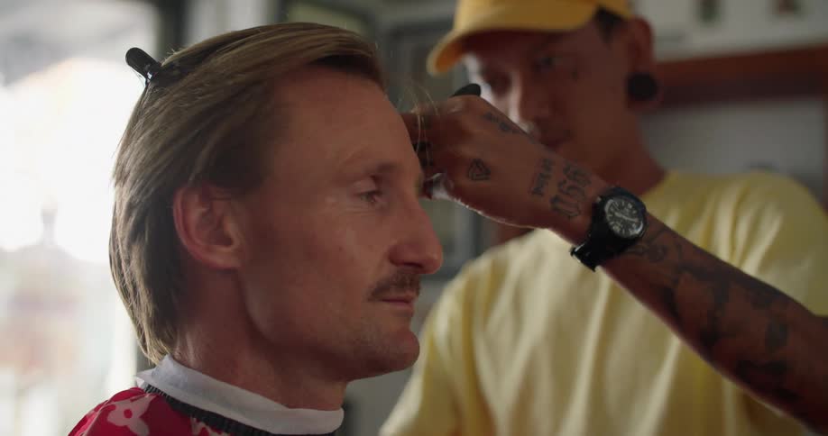 A man is sitting in a chair in a barber shop while a barber cuts his hair