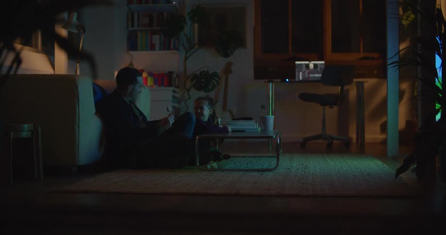 A man is sitting on the living room floor watching television with his young daughter