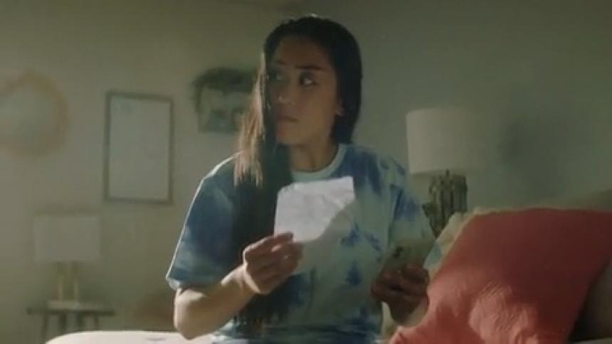 Woman with wavy hair and white and blue shirt sits on bed using smartphone and reading a paper in room