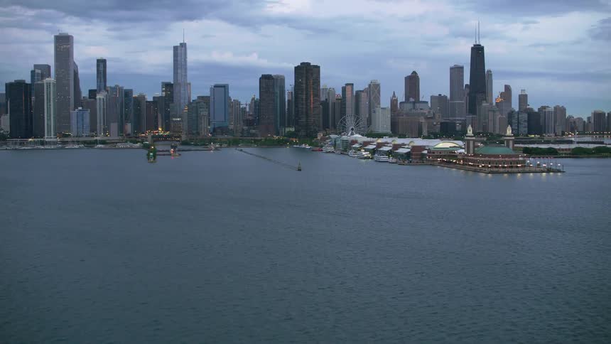Lighthouse building sits in the lake near the shoreline as city buildings stand under a cloudy sky