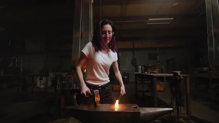 Woman wearing safety goggles heats metal in a fire then pounds the metal with a hammer