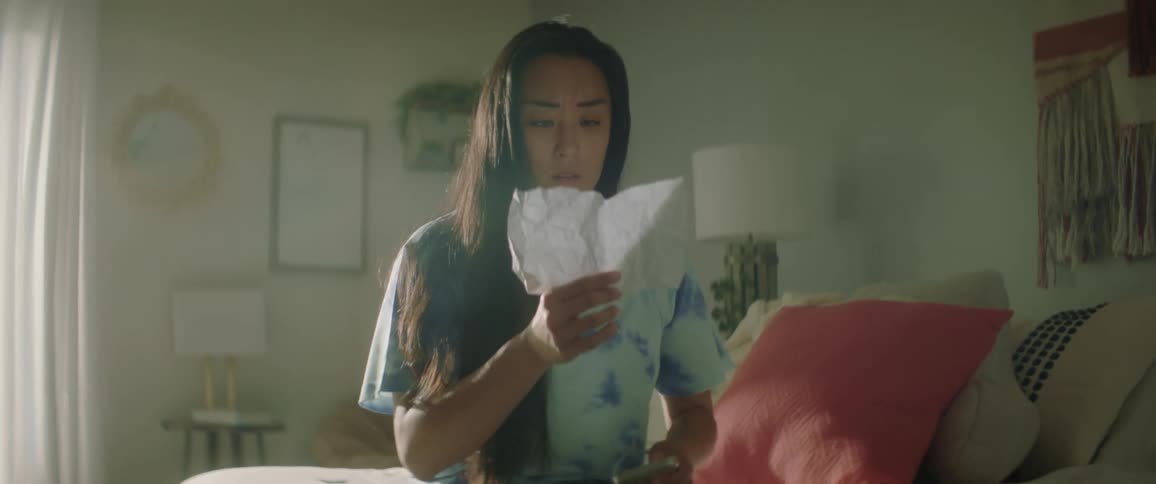 A teenage girl sits on her bed and looks at her cell phone as a note flies through the air and she reads it with an upset look on her face and flops back on her bed