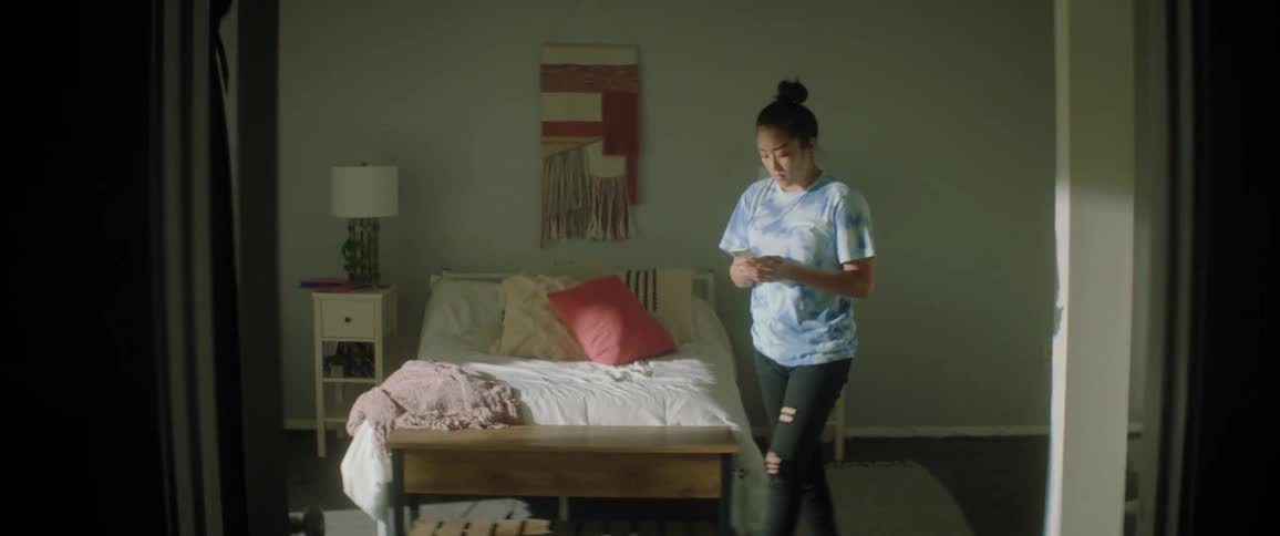 Woman with bun white blue shirt and ripped jeans stands up and walks around using smartphone in bedroom