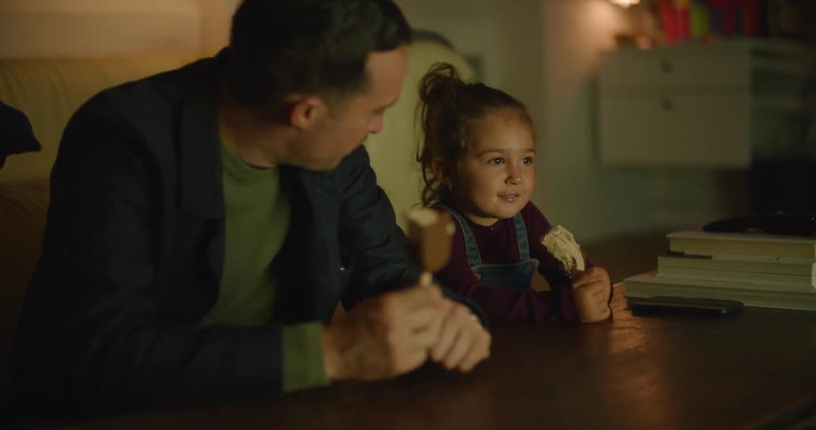 A father and young daughter eating ice cream bars while watching tv at home