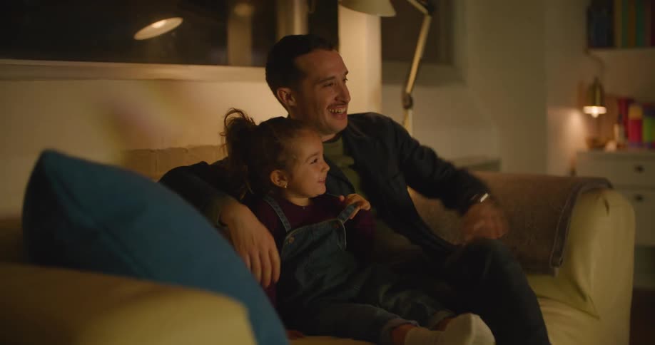 A father and young daughter smiling and laughing while watching television on a living room sofa