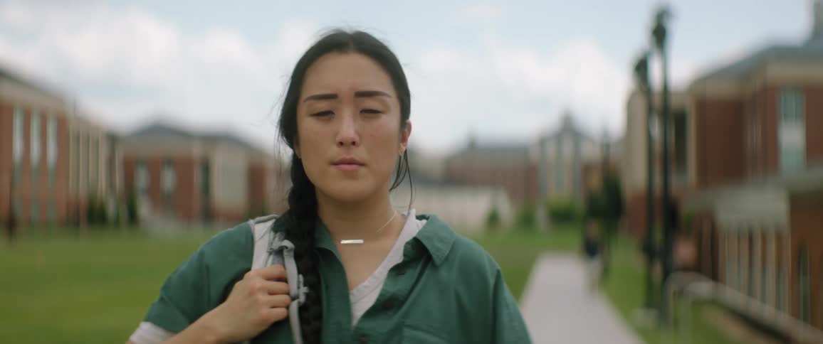 A woman is standing outside of buildings on a sidewalk holding the strap of her backpack over her shoulder