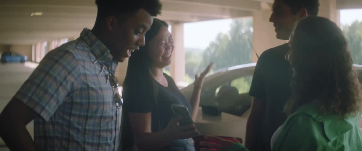 A group of friends greet each other in a parking garage and laugh while looking at a smartphone