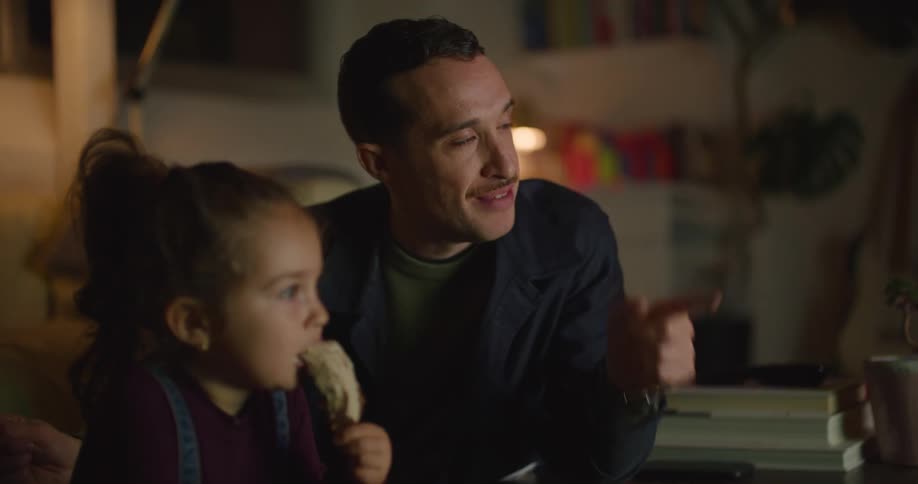 A man is leaning on a table talking to his young daughter as she eats ice cream