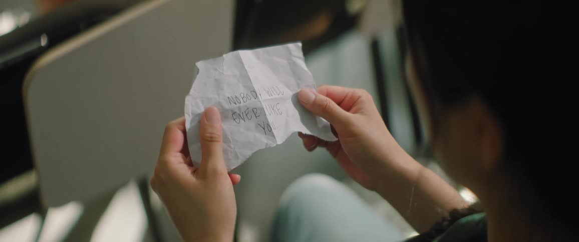 A teenager sitting in a lecture hall reads a note and looks to see who is around her