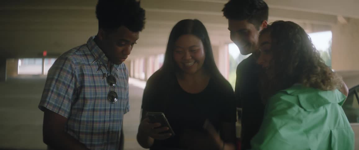 A group of teenagers in a parking garage laugh as they look at a girls cell phone