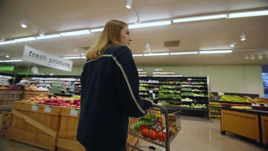 A woman is pushing a shopping cart through the produce area of a grocery store