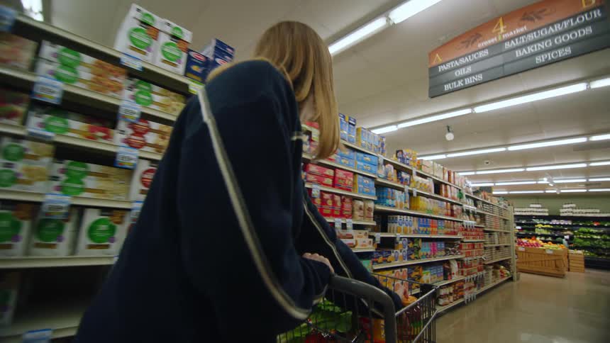 A woman is pushing a shopping cart through an aisle in a grocery store and she puts an item in her cart