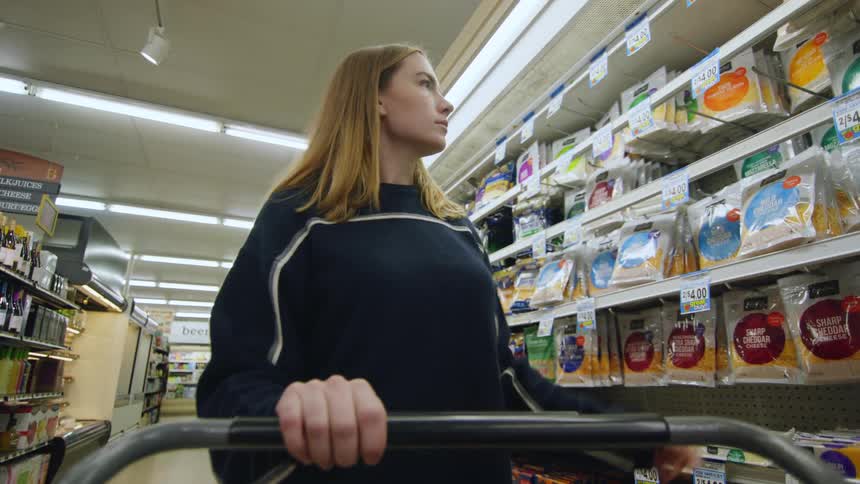 A woman putting a bag of shredded cheese in her shopping car in a grocery store