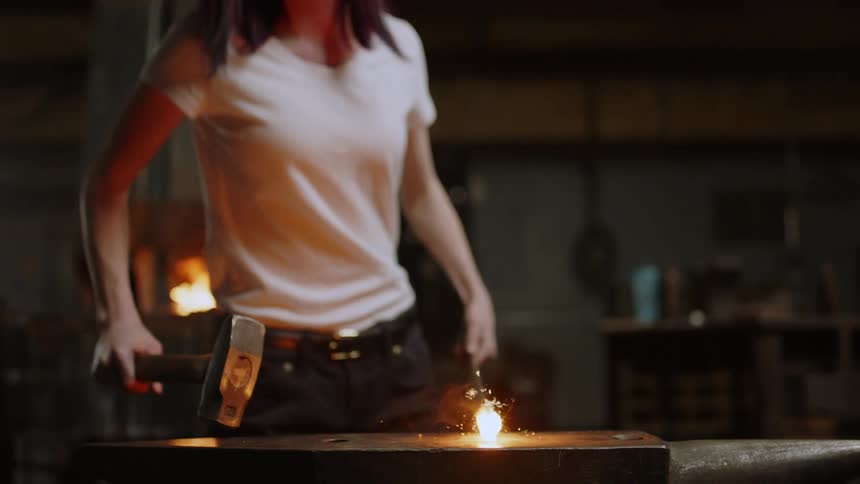 A woman removes a heated rod from a forge and places it on an anvil striking it with a hammer to shape