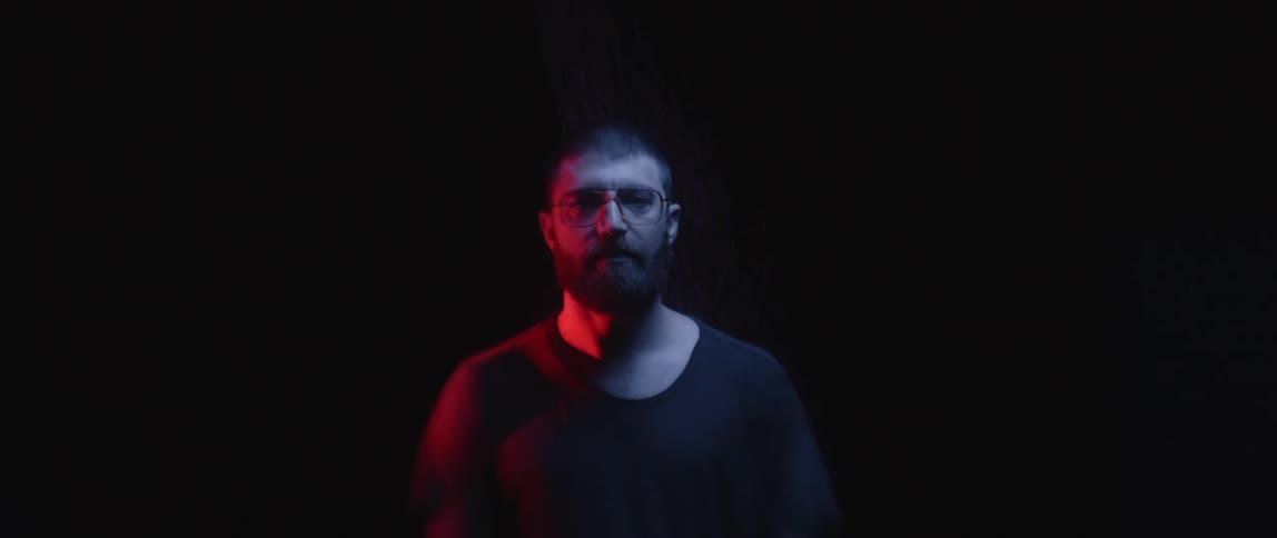 Illumination from a blinking red light is cast upon the face of a bearded man in a dark location