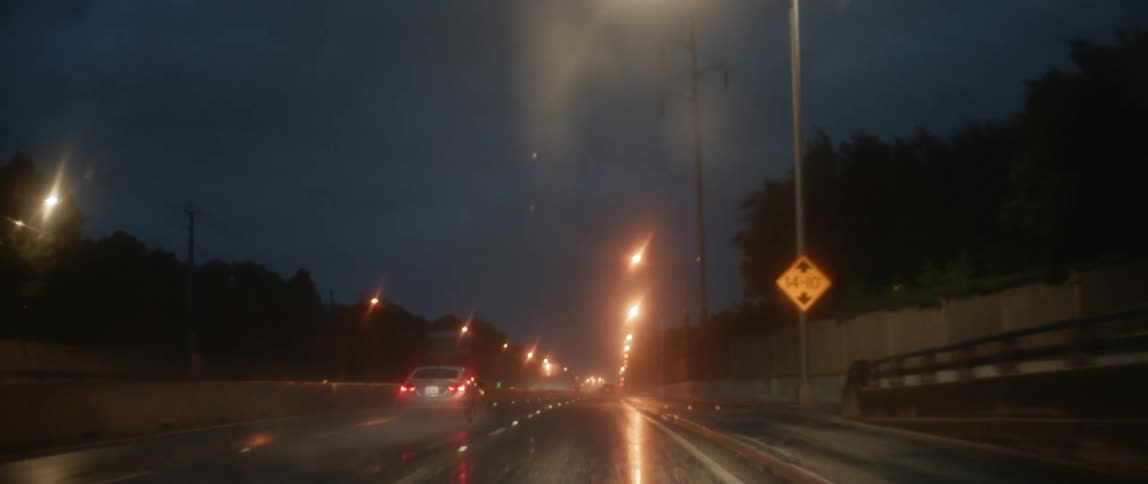 A car drives along a wide road with minimal traffic on a dark and rainy night