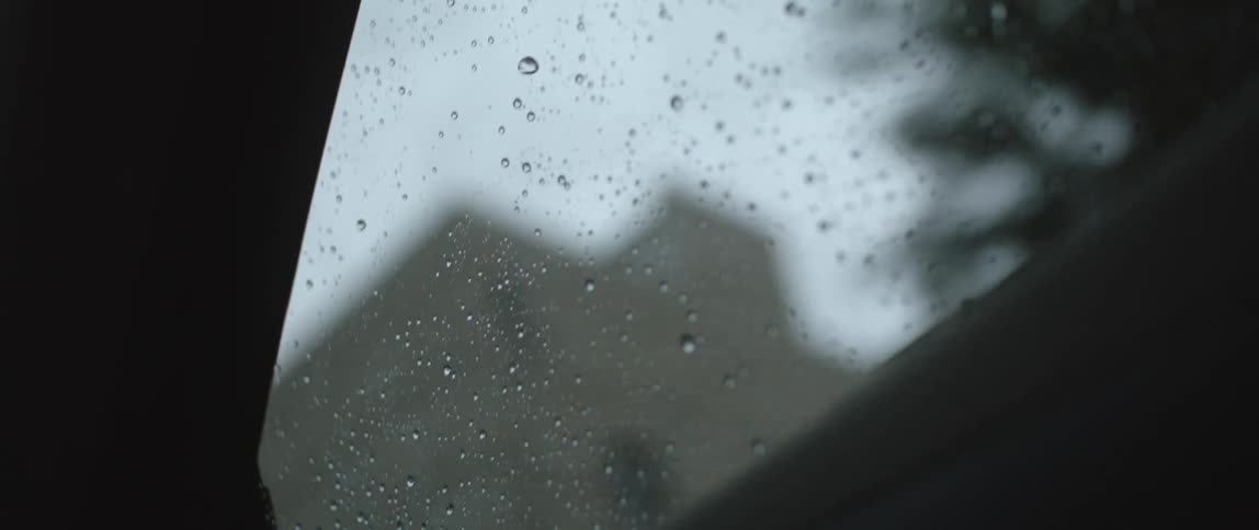 An automobile window is covered with water droplets as it travels past trees and buildings