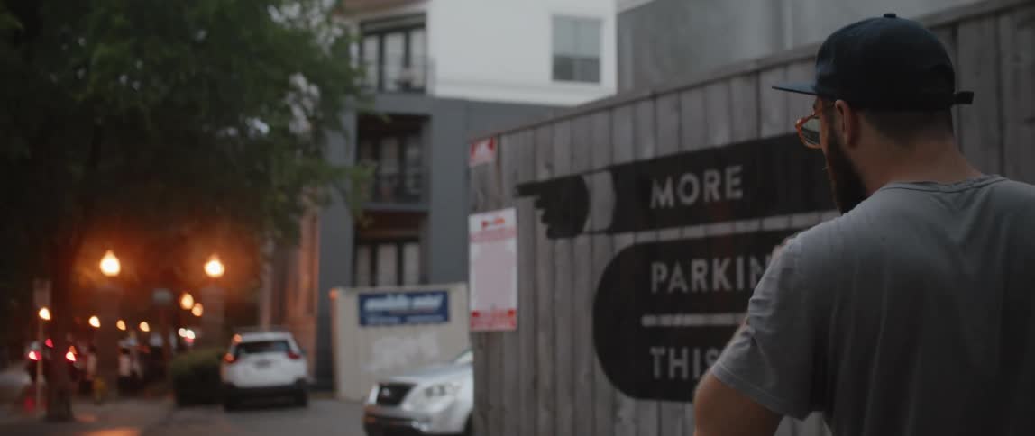 Bearded man wearing glasses and a ball cap walks past some buildings and parked cars