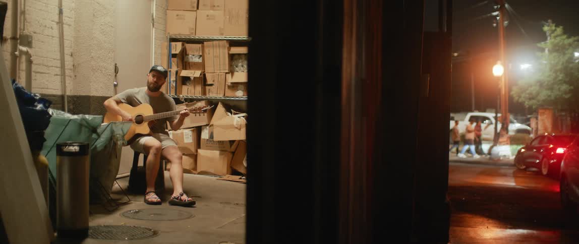 A man is sitting in a stockroom playing an acoustic guitar as cars are driving in the parking lot outside