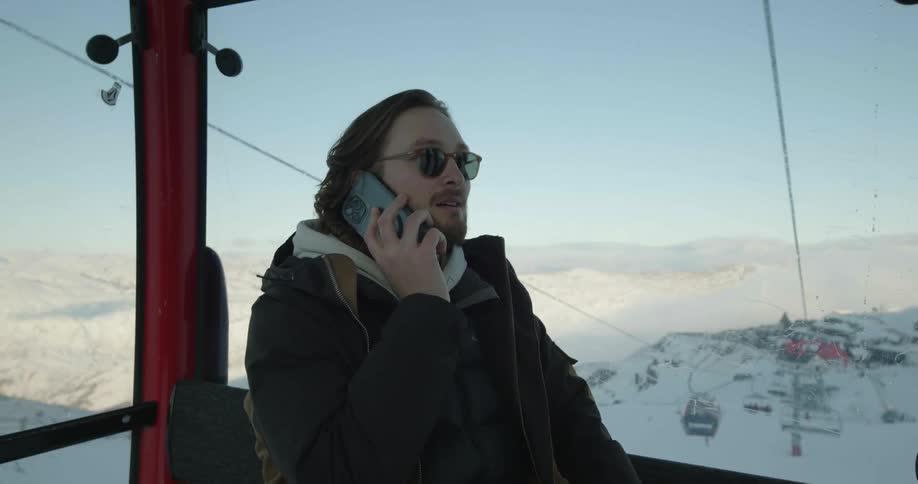 A man talks on his smartphone while riding a ski lift down a mountain slope