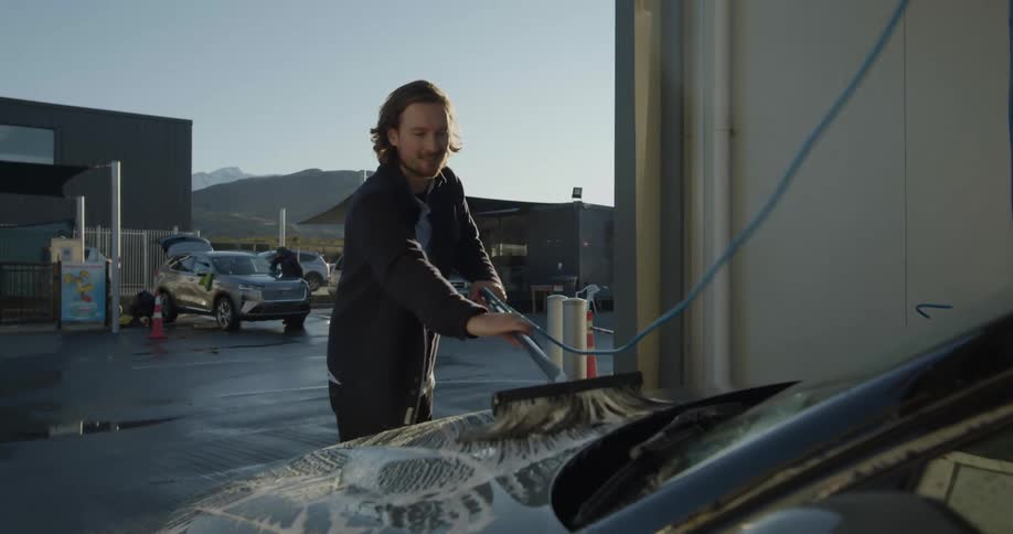 Man scrubbing the hood of his car with a soapy brush at a car wash on a sunny day