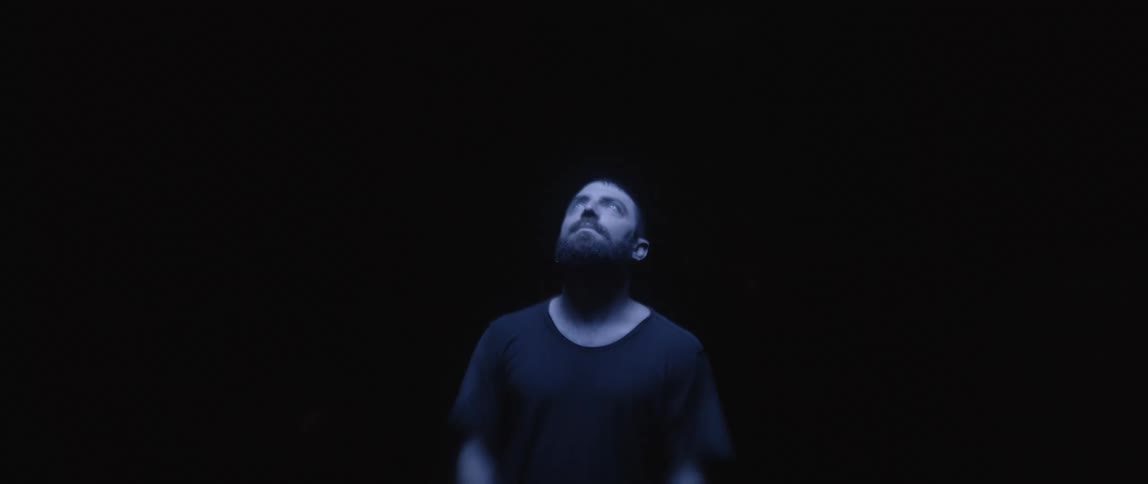 A man with a mustache and beard is standing in the darkness and he takes off his glasses