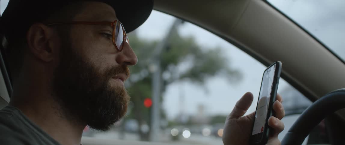 A bearded man sits in his car while looking at the screen of his phone