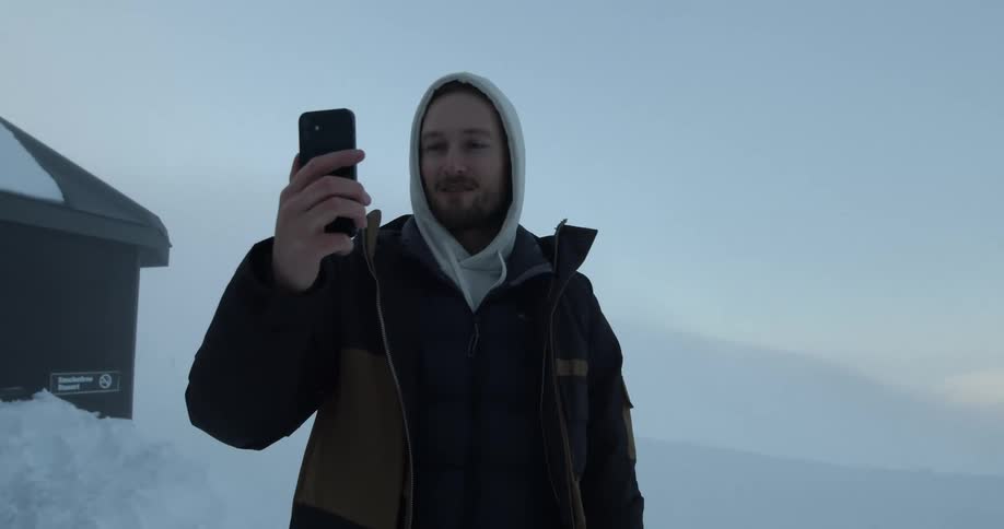 A man is standing outside in the snow with a hood on his head and holding his cell phone