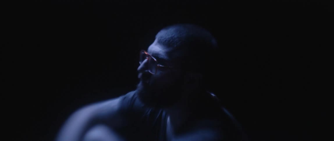 A man in glasses slowly looks upwards while sitting in a darkened place