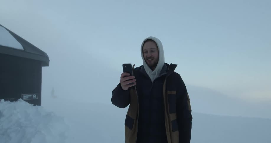 A man standing in a snowy area waves to someone his communicating with via his smart phone