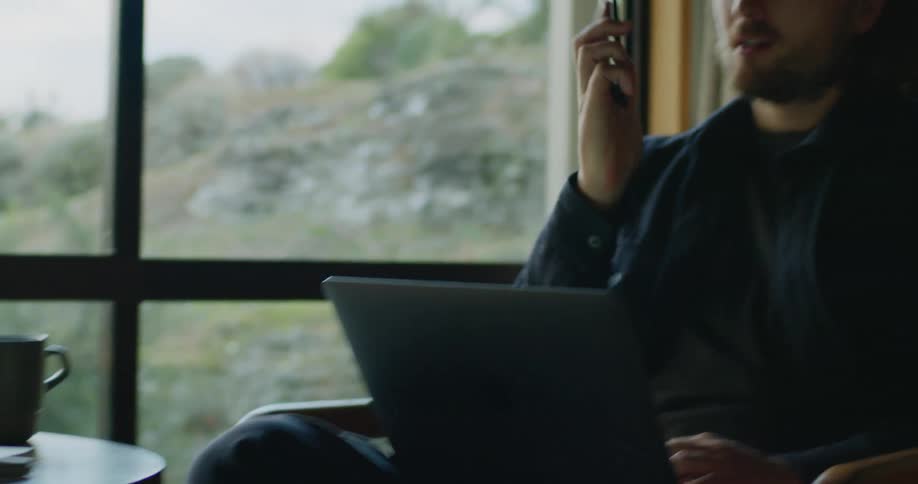 Man with a beard sits on a chair holding a laptop then picks up a smart phone from the table and begins talking