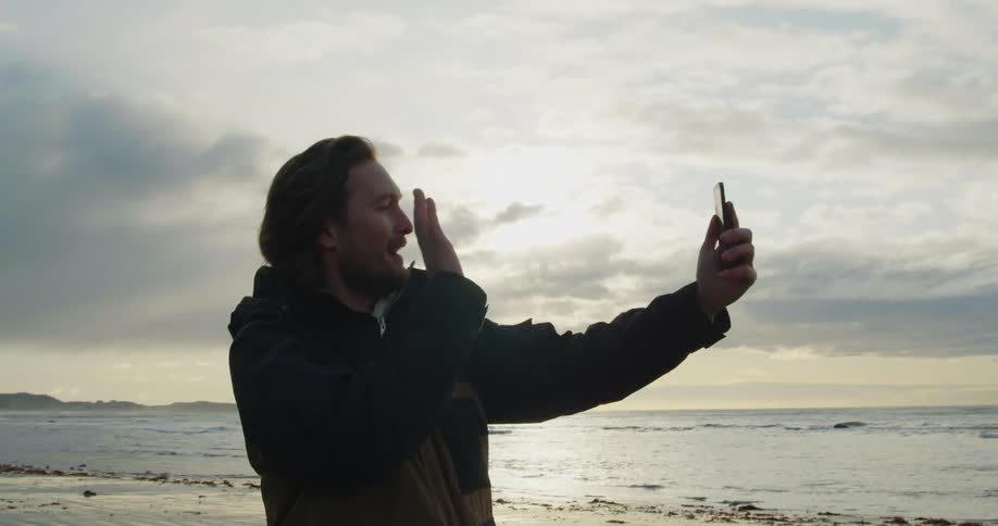 A man stands on a beach and makes a call on his cellphone and waves as he video chats