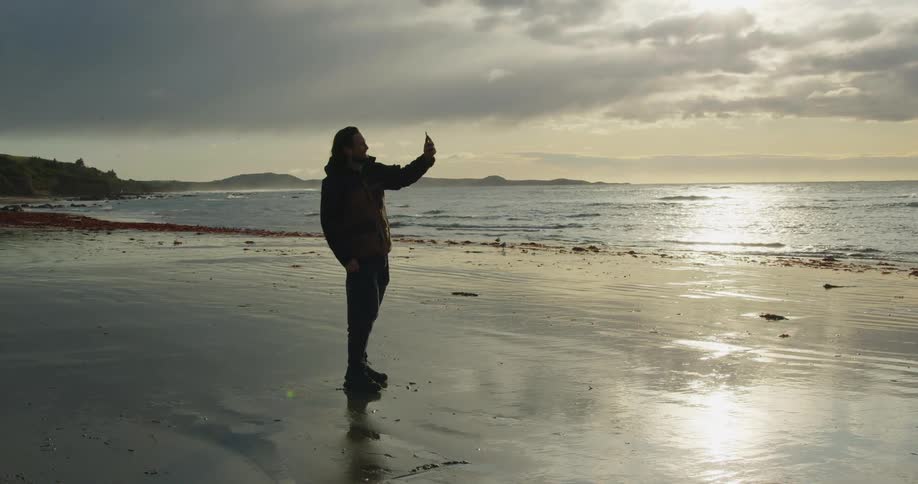 Man stands on a beach and holds up a smart phone and waves at it