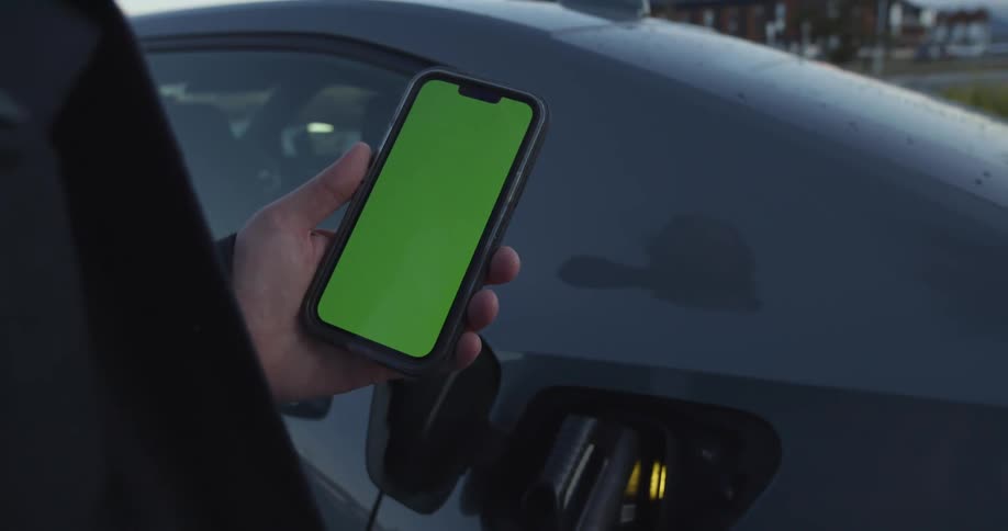 A black automobile is parked and a man reaches inside the open fuel door as he holds a cell phone with a green screen and walks away