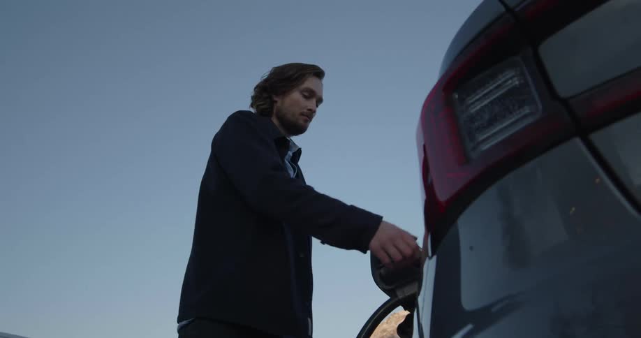 Man with long hair and beard blue shirt and naval blue jacket fills up his gray car